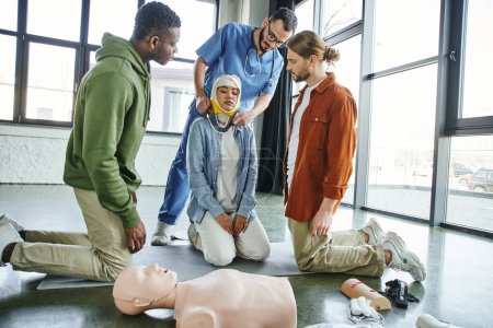 first aid training, interracial men looking at medical instructor putting neck brace on asian woman with bandaged head near CPR manikin and medical equipment, emergency situations response concept