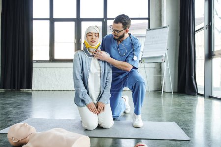 medical instructor with stethoscope, in eyeglasses and uniform, putting neck brace on asian woman with bandaged head during first aid seminar in training room, emergency situations response concept