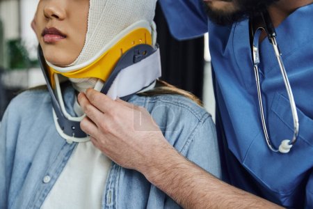 Ausgeschnittene Ansicht eines professionellen medizinischen Instruktors mit Stethoskop, der der jungen Frau mit bandagiertem Kopf eine Nackenstütze anlegt, Erste Hilfe und Notfallvorsorgekonzept