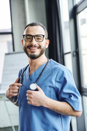 overjoyed bearded medical instructor with stethoscope on neck wearing blue uniform and smiling at camera in training room, first aid training seminar and emergency preparedness concept