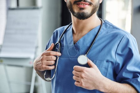 cropped view of positive bearded doctor in blue uniform touching stethoscope on neck while standing in training room, first aid training seminar and emergency preparedness concept