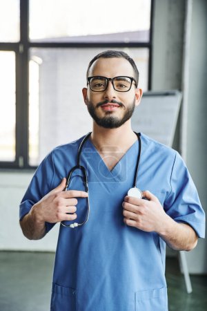fröhliche bärtige Gesundheitsfachangestellte mit strahlendem Lächeln in blauer Uniform und berührendem Stethoskop am Hals in Klinik, Erste-Hilfe-Schulung und Notfallpräventionskonzept