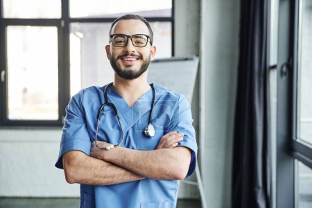 fröhlicher, bärtiger Arzt mit Brille und blauer Uniform, der mit verschränkten Armen und Stethoskop vor Kamera, Erste-Hilfe-Seminar und Notfallpräventionskonzept steht