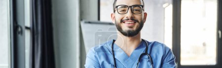 portrait of young and joyful healthcare worker with beard, eyeglasses and stethoscope looking at camera in modern clinic, first aid training seminar and emergency preparedness concept, banner