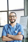 optimistic bearded paramedic in blue uniform and eyeglasses looking at camera and standing with folded arms and stethoscope near flip chart on blurred background, first aid training seminar concept magic mug #661887298