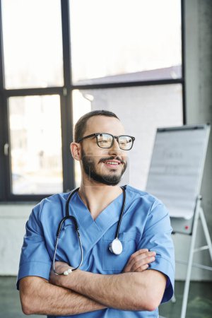Photo for Happy bearded medical instructor in eyeglasses, blue uniform and stethoscope on neck crossing arms and looking away in training room, first aid training seminar and emergency preparedness concept - Royalty Free Image