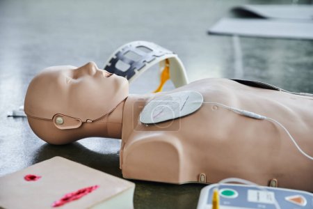 Maniquí de RCP cerca de desfibrilador externo automatizado, simulador de cuidado de heridas y soporte para el cuello sobre fondo borroso en el suelo en la sala de entrenamiento, equipo médico para entrenamiento de primeros auxilios 