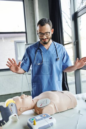 smiling bearded paramedic in blue uniform and eyeglasses gesturing near CPR manikin with defibrillator in training room, first aid hands-on learning and critical skills development concept