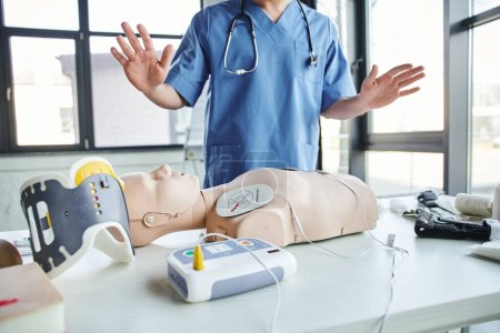 cropped view of healthcare worker in blue uniform gesturing near CPR manikin with defibrillator near tourniquets and neck brace in training room, first aid hands-on learning concept