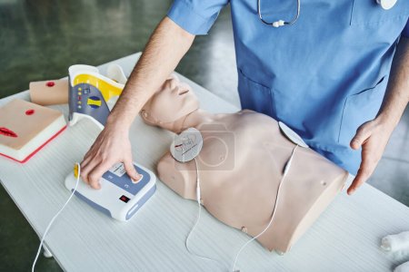 partial view of medical instructor operating automated defibrillator near CPR manikin, wound care simulators and neck brace, first aid hands-on learning and critical skills development concept