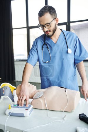 Gesundheitsfachangestellte in blauer Uniform, Stethoskop und Brille, die einen automatischen Defibrillator in der Nähe von CPR-Schaufensterpuppen bedienen, praktisches Erlernen der Ersten Hilfe und Konzept zur Entwicklung kritischer Fähigkeiten