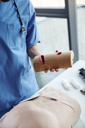 Ausgeschnittene Ansicht eines professionellen Sanitäters in blauer Uniform mit Wundversorgungssimulator in der Nähe von CPR-Schaufensterpuppe und Verbänden auf verschwommenem Hintergrund im Schulungsraum, Konzept zur Entwicklung kritischer Fähigkeiten