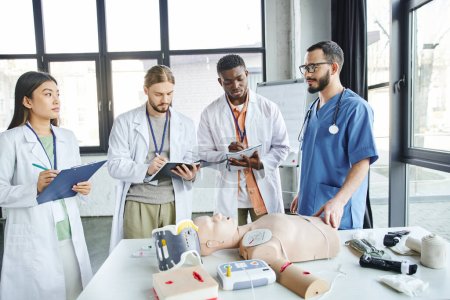 first aid seminar, diverse group of multiethnic students in white coats writing near paramedic, CPR manikin, defibrillator and medical equipment in training room, energy situations response concept puzzle 661887690