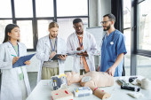 first aid seminar, diverse group of multiethnic students in white coats writing near paramedic, CPR manikin, defibrillator and medical equipment in training room, energy situations response concept puzzle #661887690