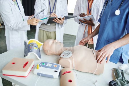 cropped view of woman in white coat pointing at CPR manikin and defibrillator near paramedic and multiethnic students writing in notebooks, life-saving skills hands-on learning concept