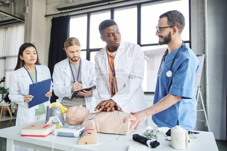 positive medical instructor looking at african american man practicing chest compressions on CPR manikin near medical equipment and multiethnic students, emergency situations response concept