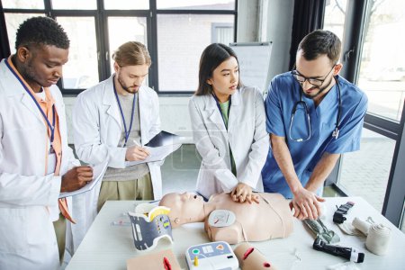 multiethnic students in white coats writing in notebooks near medical instructor assisting asian woman doing chest compressions on CPR manikin, emergency situations response concept