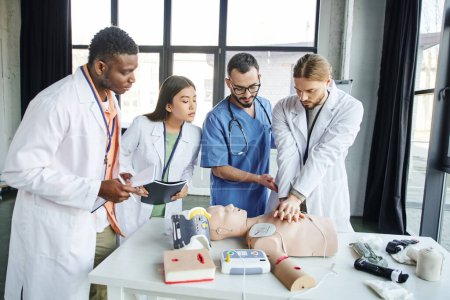 multiethnic students with notebooks looking at man doing chest compressions on CPR manikin near healthcare worker and medical equipment in training room, emergency situations response concept