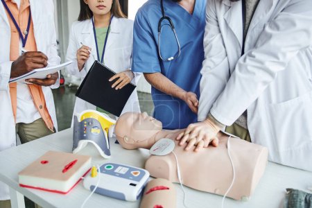 cropped view of african american man writing in notebook next to healthcare worker doing chest compressions on CPR manikin  during first aid seminar, life-saving skills hands-on learning concept