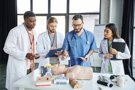 cardiac resuscitation, medical instructor holding defibrillator pads above CPR manikin near young multiethnic students in white coats, emergency situations response concept