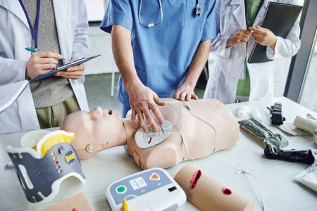 Ausgeschnittene Ansicht von Ausbildern, die Defibrillator-Pads an CPR-Schaufensterpuppen in der Nähe medizinischer Geräte anlegen und jungen Studenten in weißen Mänteln während eines Erste-Hilfe-Seminars, lebenserhaltendes praktisches Lernkonzept