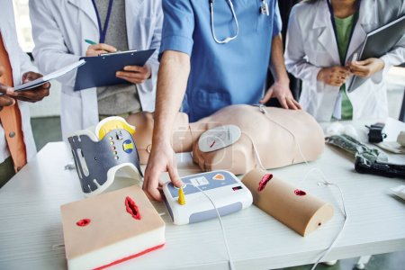 partial view of healthcare worker operating external defibrillator on CPR manikin near multicultural students with clipboard and notebooks, life-saving skills hands-on learning concept