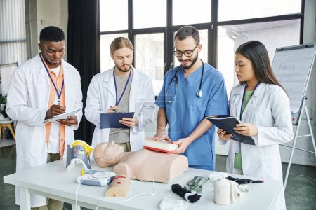 multiethnic students writing next to instructor showing wound care simulator near CPR manikin, automated defibrillator and medical equipment in training room, emergency situations response concept