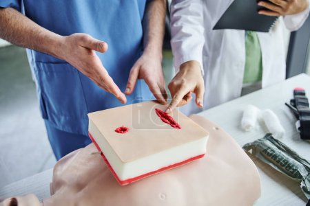 medical seminar, partial view of professional healthcare worker and student pointing at wound care simulator near CPR manikin and bandages, first aid and emergency preparedness concept