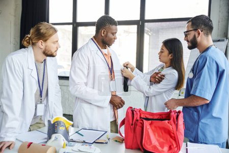 asian woman in white coat applying compressive tourniquet on arm of african american student near paramedic, first aid kit and medical equipment, life-saving skills and bleeding prevention concept