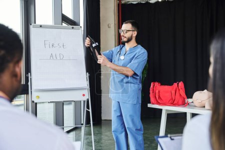 doctor in blue uniform and eyeglasses standing with compressive tourniquet near flip chart with first aid lettering and multiethnic students on blurred foreground, life-saving skills concept