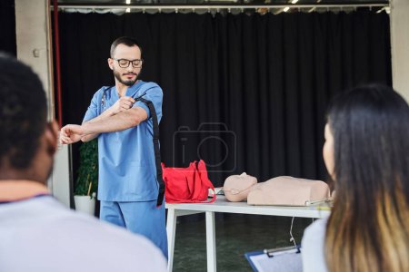 first aid seminar, medical instructor in blue uniform and eyeglasses applying compression tourniquet near interracial participants on blurred foreground, acquiring and practicing life-saving skills concept