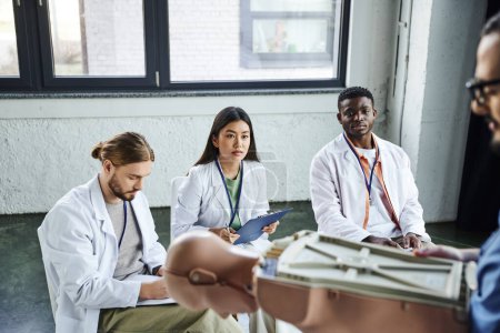 medizinische Ausbildung, vielfältige Gruppe multiethnischer Studenten in weißen Mänteln, die Sanitäter mit CPR-Schaufensterpuppe im verschwommenen Vordergrund betrachten, die lebensrettende Fähigkeiten erwerben und praktizieren