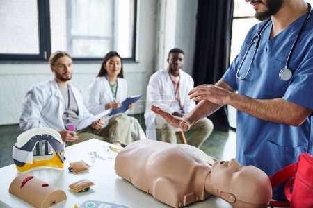 Gesundheitshelfer zeigt lebensrettende Techniken an CPR-Schaufensterpuppen in der Nähe medizinischer Geräte und eine vielfältige Gruppe von interrassischen Studenten auf verschwommenem Hintergrund, die sich lebensrettende Fähigkeiten aneignen
