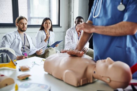 Photo for Young multiethnic students in white coats looking at paramedic showing life-saving techniques on CPR manikin on blurred foreground, acquiring and practicing life-saving skills concept - Royalty Free Image