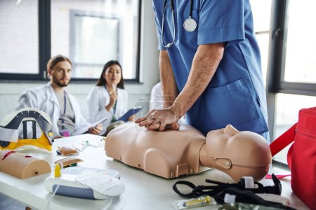 ambulancier paramédical professionnel en uniforme bleu faisant des compressions thoraciques sur mannequin de RCR près de l'équipement médical et de jeunes étudiants multiethniques sur fond flou, pratiquant concept de compétences vitales