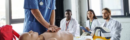 professionelle Sanitäter machen Brustkompressionen auf CPR-Schaufensterpuppe in der Nähe multiethnischer Studenten in weißen Mänteln während eines Erste-Hilfe-Seminars, erwerben und üben lebensrettende Fähigkeiten Konzept, Banner