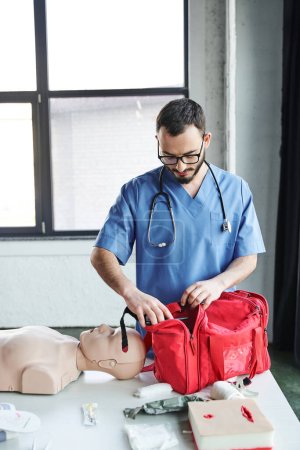 young medical instructor in eyeglasses and blue uniform preparing training room for first aid seminar and opening red bag near CPR manikin, life-saving skills development concept