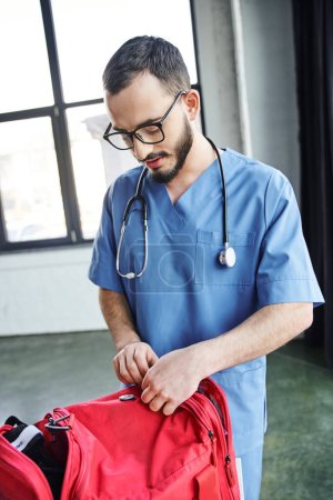 joven y barbudo trabajador de la salud en anteojos y uniforme azul desabrochando bolsa roja de primeros auxilios mientras se prepara para el seminario médico, adquiriendo el concepto de habilidades para salvar vidas