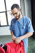 young and bearded healthcare worker in eyeglasses and blue uniform unzipping red first aid bag while preparing to medical seminar, acquiring life-saving skills concept puzzle #661888318