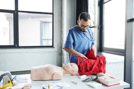young bearded doctor in blue uniform and eyeglasses unpacking red first aid bag near CPR manikin, automated defibrillator and other equipment in training room, life-saving skills development concept