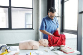 young bearded doctor in blue uniform and eyeglasses unpacking red first aid bag near CPR manikin, automated defibrillator and other equipment in training room, life-saving skills development concept Tank Top #661888324