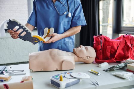 partial view of healthcare worker in blue uniform holding neck brace near CPR manikin, defibrillator, first aid kit and medical equipment, life-saving skills development concept