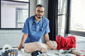 bearded paramedic in eyeglasses and blue uniform looking at camera near CPR manikin, red first aid bag, defibrillator and neck brace in training room, life-saving skills development concept Longsleeve T-shirt #661888348