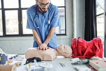 young professional paramedic practicing chest compressions on CPR manikin near red first aid bag, automated defibrillator and medical devices in training room, life-saving skills development concept