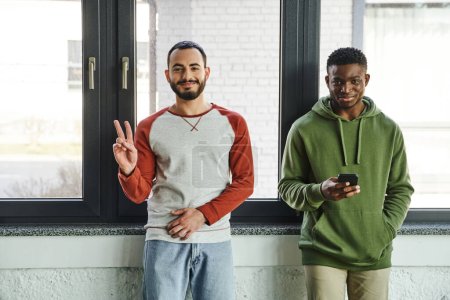 young interracial business colleagues in stylish casual clothes looking at camera in modern office, cheerful bearded man showing victory sign near african american man networking on mobile phone