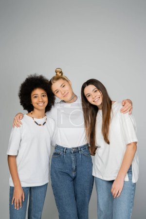 Stylish and cheerful interracial teenage girls in white t-shirts and blue jeans hugging and looking at camera while standing isolated on grey, teenage friends having fun together
