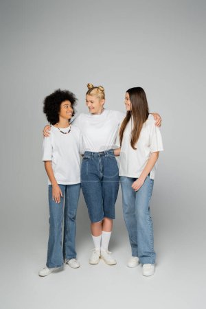 Full length of cheerful blonde teen girl hugging positive multiethnic girlfriends in white t-shirts and stylish blue jeans while standing on grey background, teenage friends having fun together