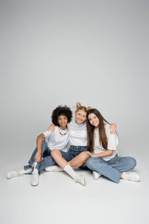 Téléchargez les photos : Pleine longueur de souriantes amies adolescentes interracial en t-shirts blancs et jeans bleus regardant la caméra tout en étant assis ensemble sur fond gris, concept de modèles adolescentes multiethniques - en image libre de droit