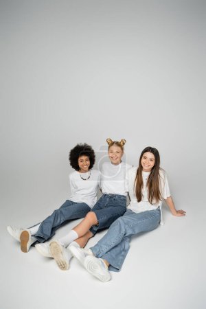 Foto de Alegre y multiétnico adolescentes novias en jeans de moda y camisetas blancas mirando a la cámara mientras se sienta y posando sobre fondo gris, multiétnico modelo adolescente concepto - Imagen libre de derechos