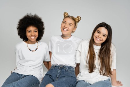 Cheerful teen and multiethnic girlfriends in white t-shirt and blue jeans sitting next to each other and looking at camera while spending time on grey background, multiethnic teen models concept